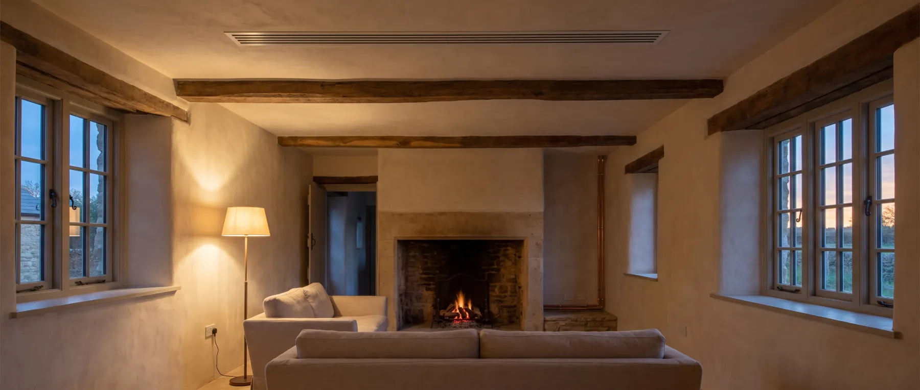 A stone farmhouse living room at dusk with a fireplace and a slim linear air grille in the ceiling