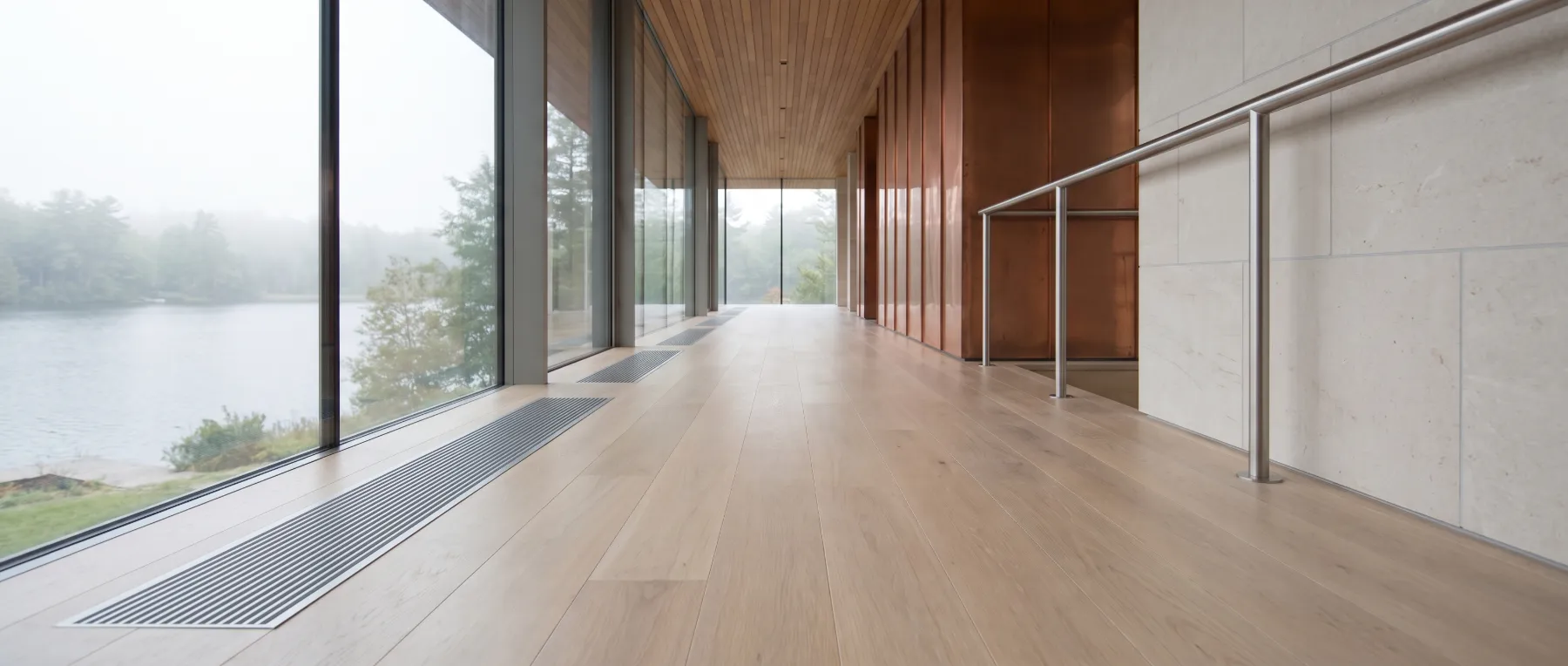 A lake house hallway with an oak floor and slim steel floor grilles along a glass wall
